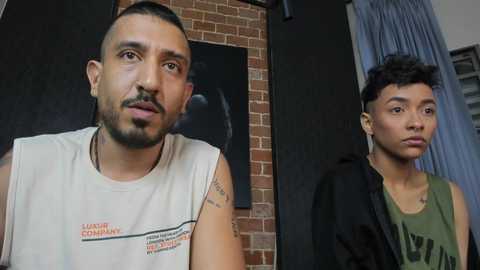 Video of a man with a beard in a white tank top and a young man in a green shirt and jacket, standing in front of a brick wall with a black framed painting.