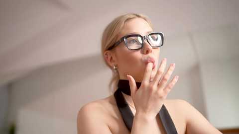 Video of a blonde woman with glasses, wearing a black dress, holding a finger to her lips in a thoughtful pose. The background is blurred, featuring a beige wall and a ceiling.