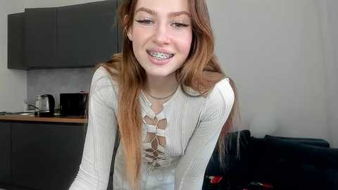 Media: Video of a young woman with long auburn hair, wearing a light sweater with a geometric pattern, grinning with braces, in a modern kitchen setting.