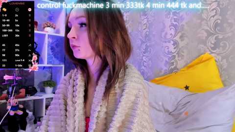 Video of a young Caucasian woman with shoulder-length brown hair, wearing a fluffy beige robe, sitting on a bed. Background includes a yellow pillow and a gray, patterned wall. Text overlay reads \"control fuckmachine 3 min 34 sec 4 min 41 sec...\