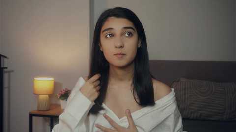 Media: Video of a young woman with medium brown skin and long black hair, wearing a white off-shoulder shirt, sitting on a bed with a lamp on a nightstand.