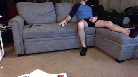 Media: Video of a man in a blue and black shirt and black shorts, playing a video game on a grey couch in a cluttered living room.