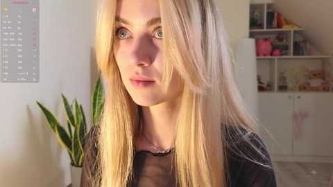 Media: Video of a blonde woman with blue eyes, wearing a black sheer top, standing in a brightly lit room with shelves, plants, and a calendar on the wall.