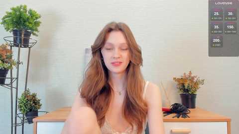 Media: Video of a young woman with long, wavy brown hair, wearing a white lace bralette, sitting on a wooden desk. Background features a white brick wall, potted plants, and a digital clock displaying time and weather.