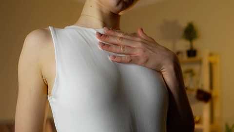 Media: Video of a woman with fair skin, wearing a white sleeveless top, standing indoors. She gently touches her chest with her left hand, her right arm partially visible. Background features a green plant and a bookshelf.