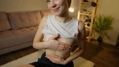 Media: Video of a smiling woman with light skin, small breasts, wearing a white crop top, revealing her bare chest, standing indoors.