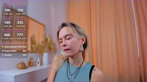 Media: Video of a fair-skinned, blonde woman with long hair, wearing a blue sleeveless top, standing in a well-lit room with beige curtains. The background includes a white dresser with gold figurines and a plant.