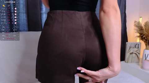 Video of a woman's lower back and hips in a black top and brown suede skirt, holding a white purse. Background includes a calendar, potted plants, and a candle.