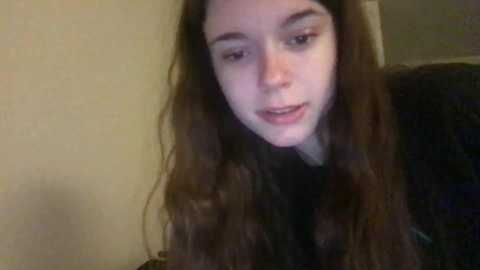 Media: Video of a young Caucasian woman with long, wavy brown hair, pale skin, and dark eyebrows, wearing a black top, looking down in a dimly lit room with beige walls.