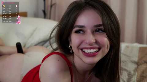 Media: A video of a young woman with long brown hair, fair skin, and a bright smile, wearing a red dress, lying on a bed with beige sheets, and a partially visible person in the background.