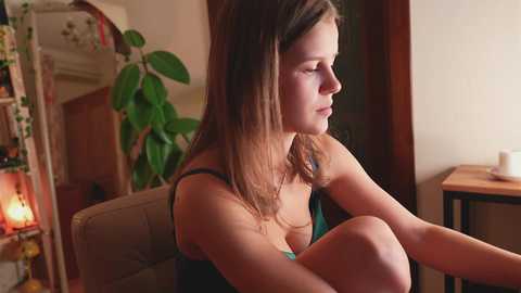 Video of a young Caucasian woman with long brown hair, wearing a green tank top, sitting pensively indoors, with a potted plant and warm lighting in the background.