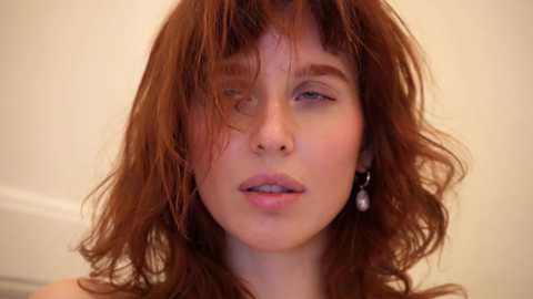 Media: Video of a young woman with wavy, shoulder-length auburn hair, pale skin, and full lips. She wears small, dangling earrings. The background is a plain, off-white wall.