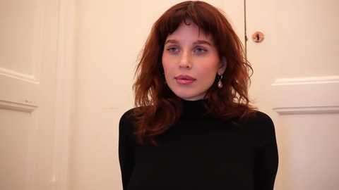 Media: Video of a Caucasian woman with wavy, shoulder-length auburn hair, wearing a black turtleneck sweater, standing in front of white paneled doors. She has fair skin and a neutral expression.