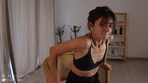 Media: Video of a slender, young Asian woman with medium-length dark hair, wearing a black bra and pants, leaning on a wicker chair in a minimalist room with light wooden floors and beige curtains.