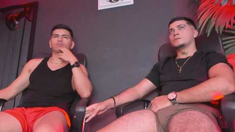 Media: Video of two young, fit men with short hair, wearing black t-shirts and red shorts, seated side by side in black leather chairs against a dark wall.