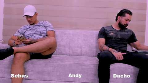 Video of two men, Seb\u00e1s and Andy, sitting side-by-side on a light-gray couch. Seb\u00e1s, in a white cap and gray patterned shirt, plays with his phone. Andy, in a black T-shirt and jeans, looks downcast. Background shows a beige wall.