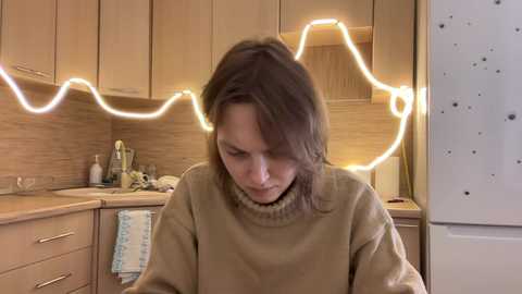 Video of a Caucasian woman with shoulder-length brown hair, wearing a beige sweater, focused on a kitchen counter with wooden cabinets, a white fridge, and a string of glowing lights forming a heart shape.