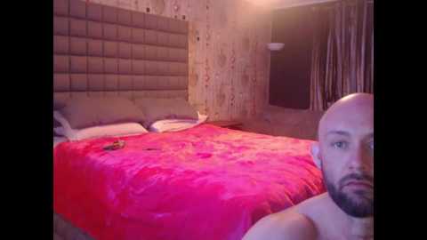 Media: Video of a dimly lit, sparsely furnished bedroom with a bald, bearded man in the foreground. The bed has a bright pink floral blanket and beige headboard.