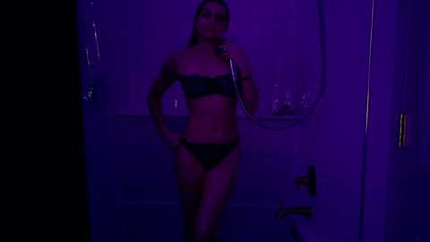 Media: Video of a slim, dark-haired woman in black lingerie, holding a microphone, in a dimly lit bathroom with purple lighting, suggesting a sultry, intimate setting.