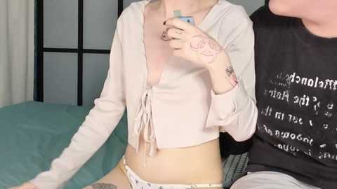 Media: Video of a fair-skinned, slender woman with tattoos, wearing a pink cropped top and white panties, sitting on a bed. A man in a black shirt with white text is partially visible.