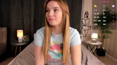 Media: Video of a young woman with long blonde hair, wearing a colorful tie-dye T-shirt, sitting on a bed in a dimly lit room with a laptop displaying a dating app screen.