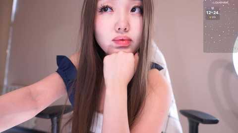 Video of a young Asian woman with long brown hair, pale skin, and a serious expression, resting her chin on her hand in a beige office setting with a calendar in the background.