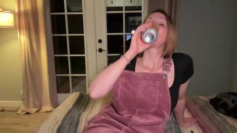 A video of a woman with light skin, blonde hair, and wearing a black shirt and pink overalls, drinking from a silver can, sitting on a striped bed in a dimly lit room with beige walls and wooden floors.