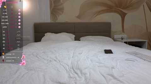 A video of a modern bedroom with a large bed, white sheets, and a grey headboard. A smartphone lies on the bed, and a white nightstand holds a lamp. A mural of abstract tree branches adorns the wall.