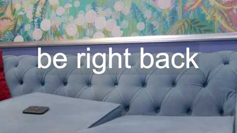 A video of a modern, light blue tufted leather bench seat in a colorful, bohemian-themed restaurant. The background features a mural with vibrant floral and leaf patterns. The text overlay reads, \"be right back.\