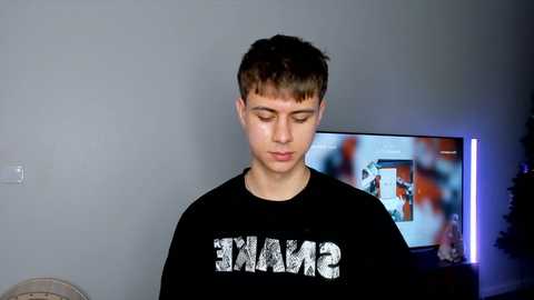 Media: Video of a young, fair-skinned, short-haired boy in a black shirt, indoors, looking down, with a TV displaying an action movie behind him.