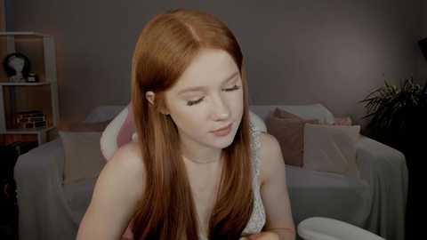 Video of a fair-skinned young woman with long, straight red hair, wearing a white lace sleeveless top, sitting in a dimly lit living room with a gray couch, a bookshelf, and a potted plant in the background.