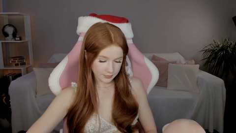 A video of a fair-skinned, red-haired woman with long hair, wearing a pink bunny headband and a white and pink dress, sitting on a couch in a dimly lit room.