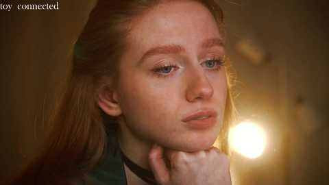 Media: Video of a pale-skinned young woman with long, light brown hair, wearing a dark green top, gazing thoughtfully, with a soft, warm background featuring a bright, circular light source.