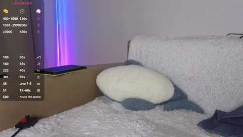 Video of a plush toy resembling a white and blue dog with a fluffy blanket, placed on a light-colored rug, against a plain white wall. A black smartphone and a purple LED strip light are visible in the background.