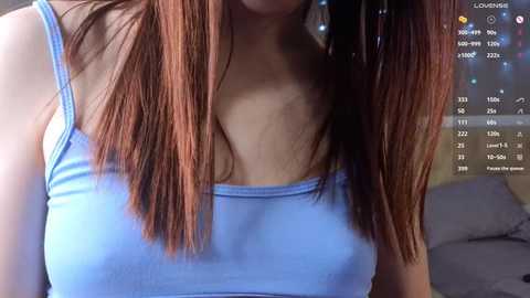 Video of a young woman with long, straight brown hair in pigtails, wearing a light blue tank top that reveals her medium-sized breasts, in a dimly lit room with a computer screen showing a date and time in the background.