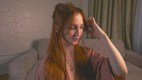 Video of a fair-skinned, red-haired woman in a light pink robe, adjusting her hair in a dimly lit, modern living room with white patterned wallpaper and beige furniture.