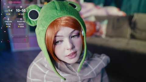 Media: Video of a young woman with pale skin, bright red hair, and wearing a green frog costume, indoors with blurred background.