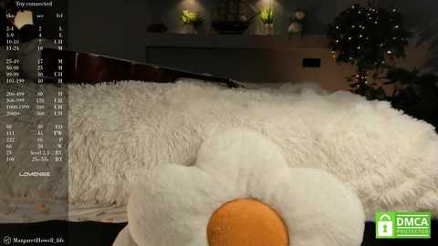 Media: Video of a plush, white, fluffy pillow resembling a flower with an orange center, set on a bed, against a dimly lit room with potted plants and a wooden headboard.