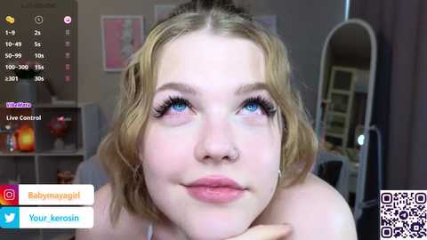 A video of a young woman with fair skin, blonde hair, and blue eyes, wearing light makeup, sitting in a cozy room with a mirror and colorful decor.