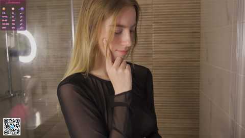 Video of a young Caucasian woman with long, straight blonde hair, wearing a black sheer top, standing in a dimly lit bathroom with beige tiled walls and blinds. She appears introspective, with her hand on her chin.