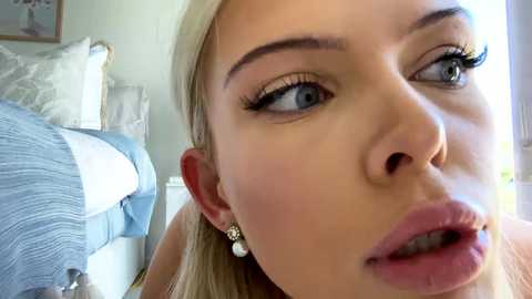 Media: Video of a close-up of a young blonde woman with fair skin, blue eyes, and full lips, wearing gold earrings, looking slightly surprised or amused, in a bedroom with a white bed and blue bedding.