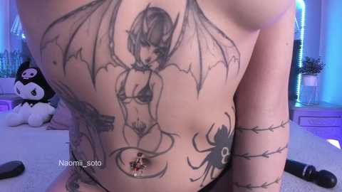 Video of a pale-skinned, tattooed woman with a demonic winged figure, a spider, and a small red heart on her abdomen, sitting on a bed with a plush toy panda and a pink nightstand in the background.