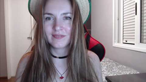 Media: Video of a fair-skinned, blonde woman with a straw hat, wearing a black choker necklace, sitting indoors near a window with white blinds.