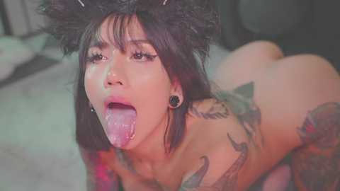 Media: Video of a topless woman with dark hair, wearing a black furry hat, licking her tongue, tattoos on her arms, in a blurry, dimly lit room.