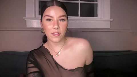 Media: Video of a fair-skinned woman with dark hair in a low ponytail, wearing a sheer black off-shoulder top and gold hoop earrings, sitting indoors with a white-framed window and dark couch in the background.