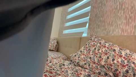 A video captures a bed with floral-patterned bedding, beige headboard, and a partially visible window with horizontal blinds. The image is taken from a low angle, emphasizing the bed's cozy appearance.