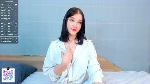 Video of a fair-skinned woman with long black hair, wearing a white button-down shirt, seated on a bed with white sheets in a minimalist room.