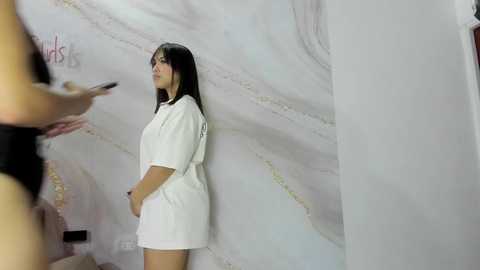 Video of a young woman with long black hair, wearing a white robe, standing against a marble wall. She looks at a smartphone held by another person out of frame.