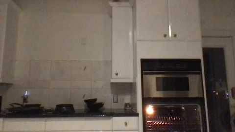 Media: Video of a dimly lit, minimalist kitchen with white cabinets, stainless steel appliances, and beige tiled backsplash. A microwave oven and a stovetop are visible, along with a bowl on the countertop. The room has a contemporary, clean look.