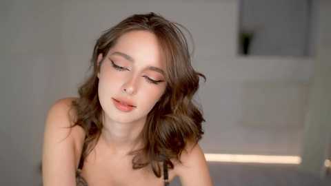 Media: A video of a young woman with wavy brown hair, wearing a black bra, closing her eyes with a soft smile, in a softly lit, minimalist room.